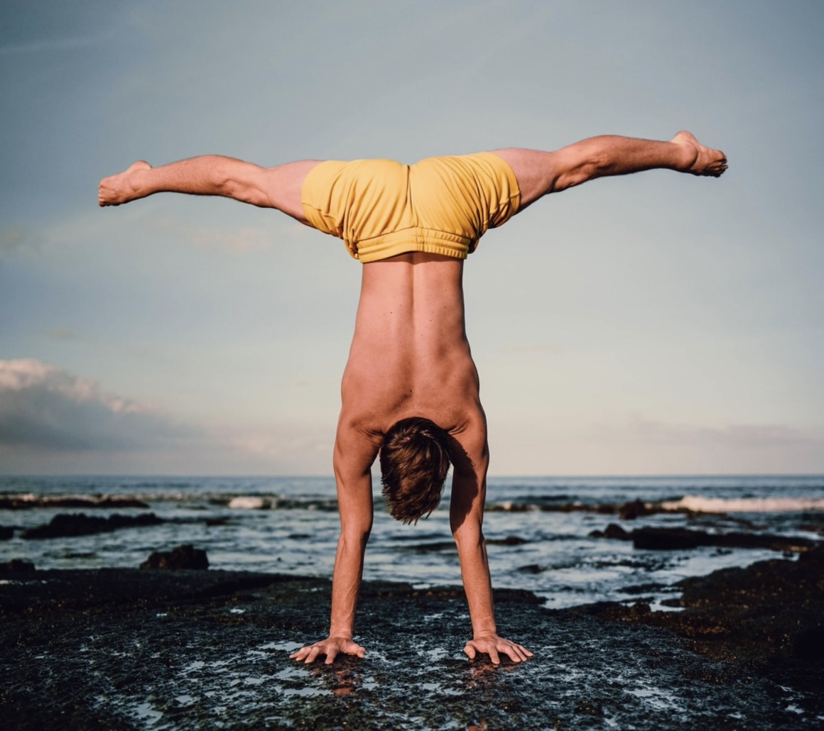 A straddle handstand held on a rocky shoreline at sunset